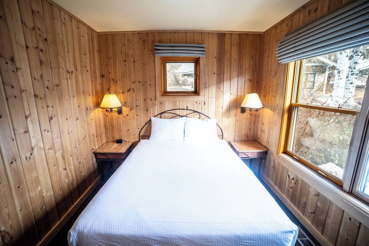 A wooden-paneled room with a bed, two bedside lamps, a window, and a small skylight.