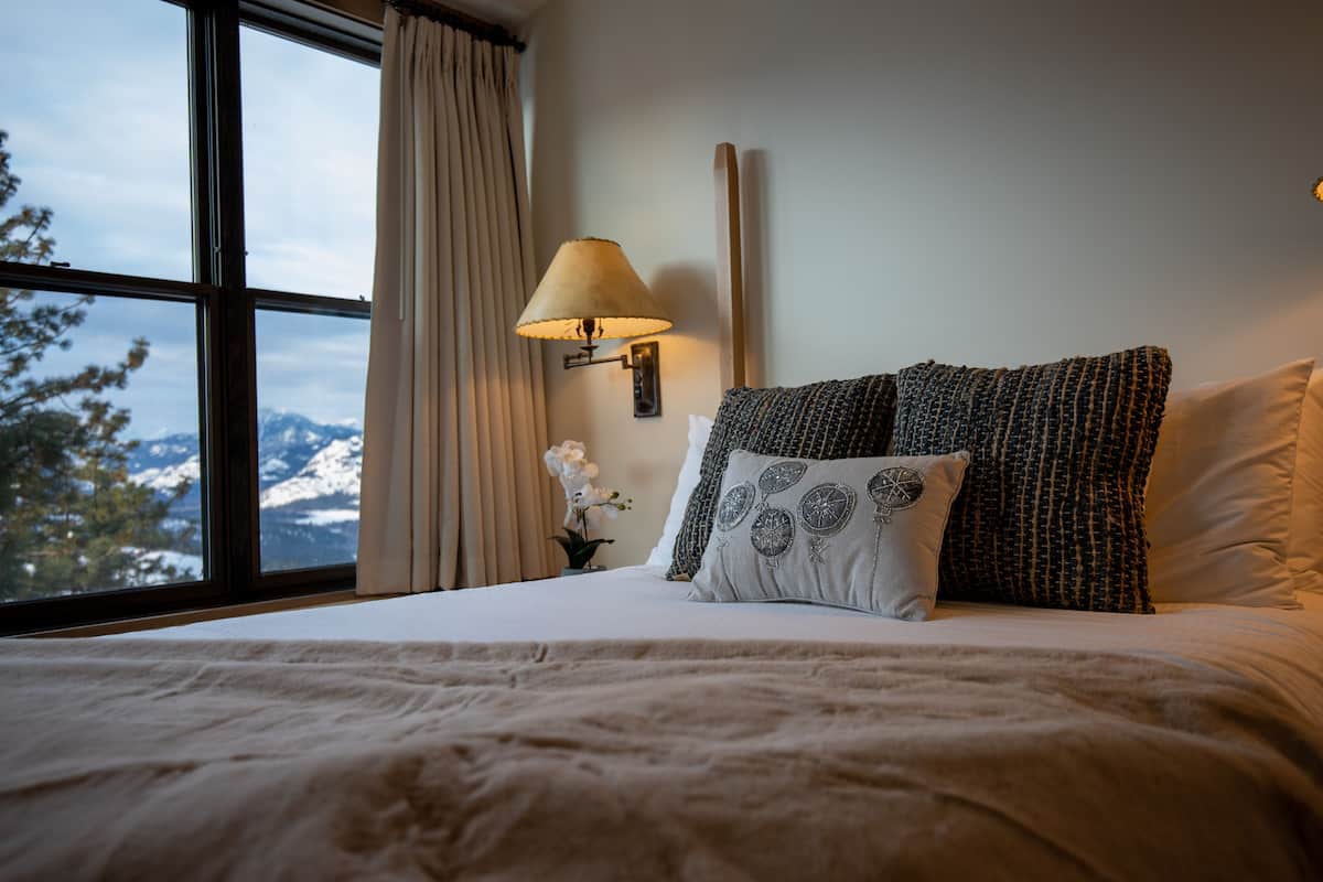 A bedroom with a large window, a bed with patterned pillows, a bedside lamp, and a view of snow-capped mountains.