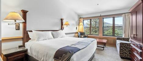 A hotel room with a large bed, two bedside lamps, a window with a view, a desk, and a chair.