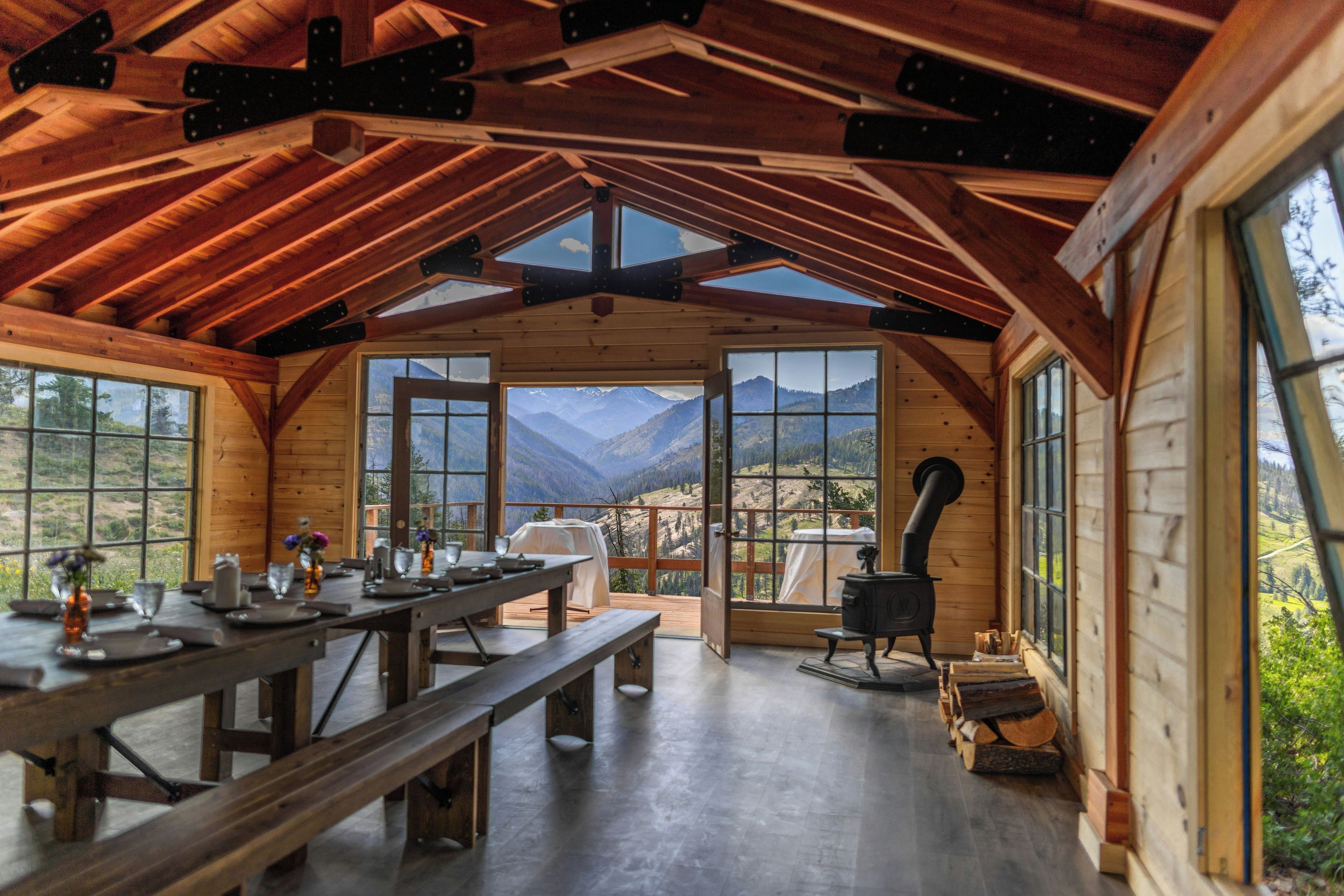 A wooden cabin interior with a dining area, large windows, and a wood stove.
