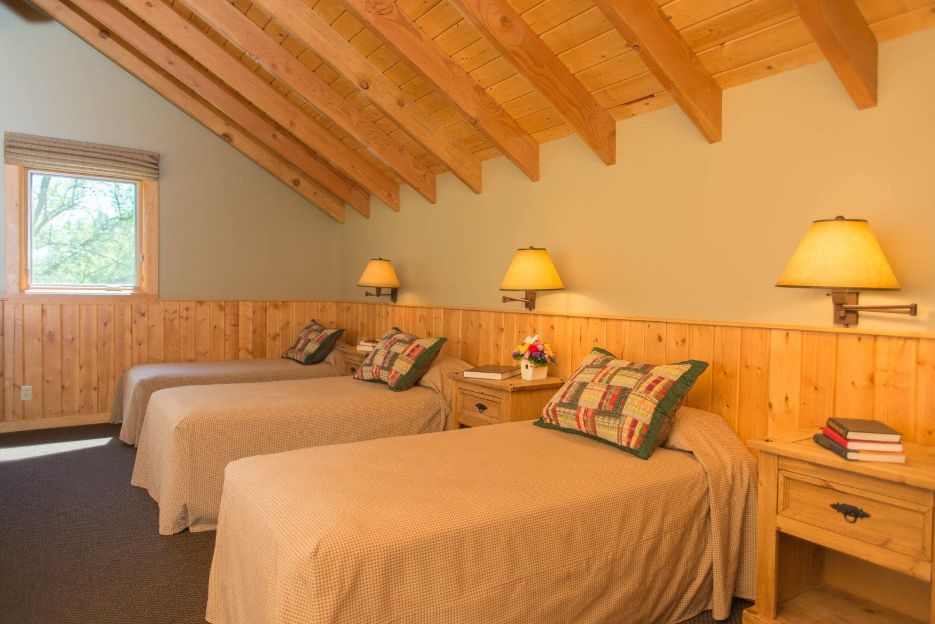 A room with two beds, wooden walls, a sloped ceiling, and wall-mounted lamps.