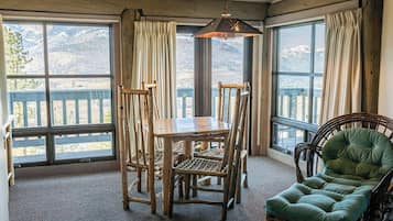 Une pièce comprenant une table et des chaises en bois, un fauteuil recouvert d’un tissu vert et de grandes fenêtres avec des rideaux.