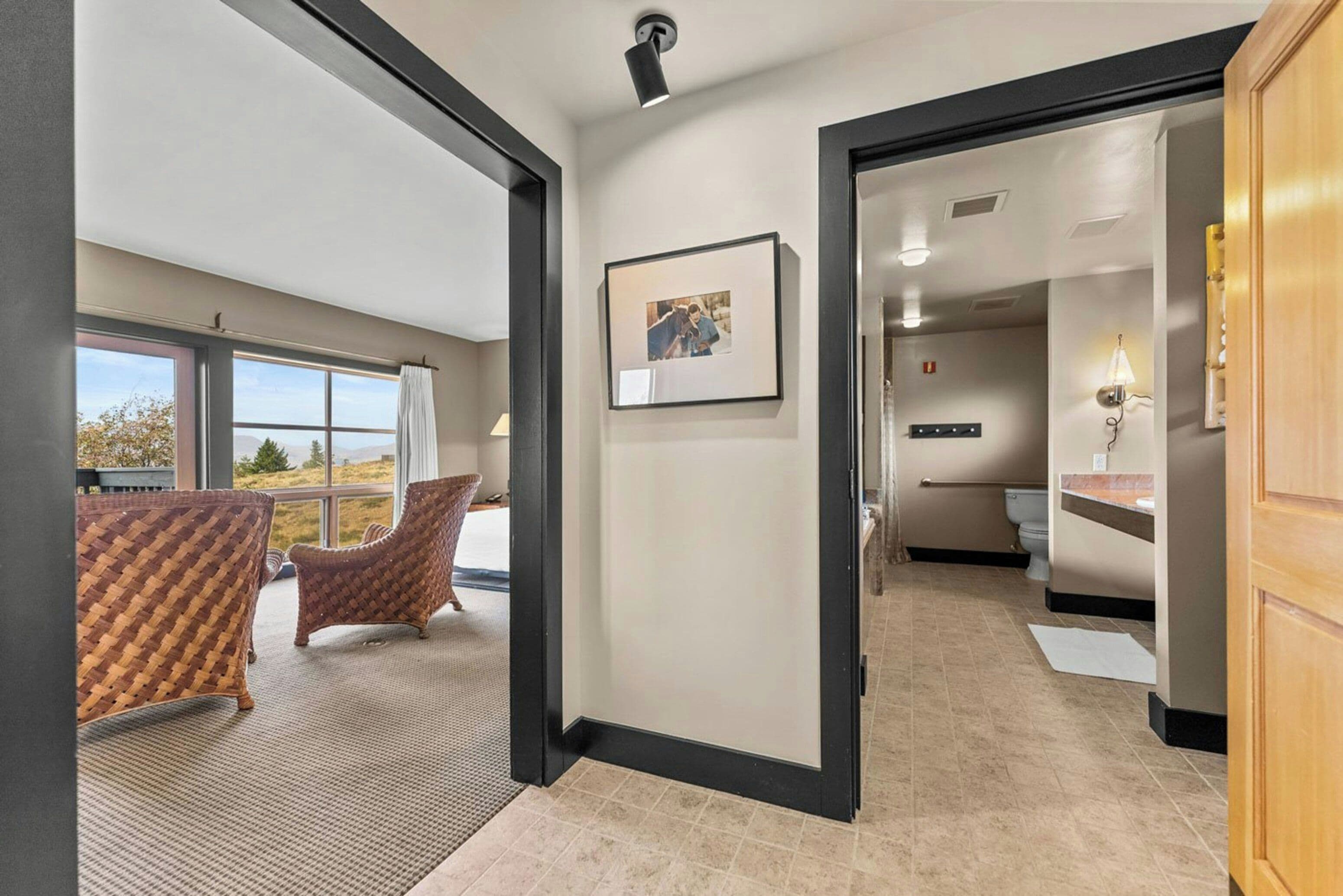 A hotel room with a large window, a framed picture on the wall, and a wicker chair.