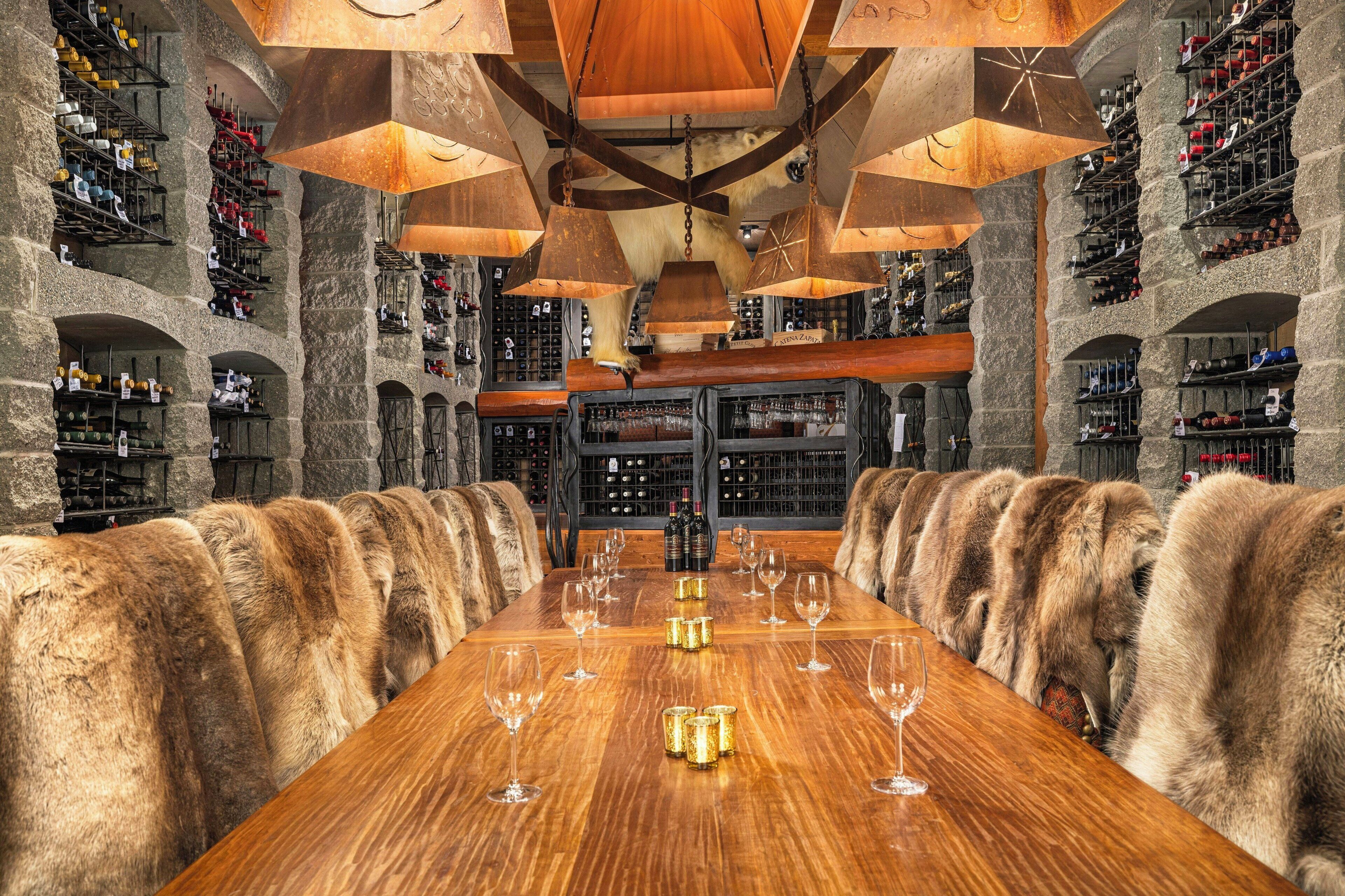 Une cave à vin spacieuse, avec une longue table en bois dressée pour le repas, entourée de murs en pierre et de présentoirs métalliques remplis de bouteilles de vin.