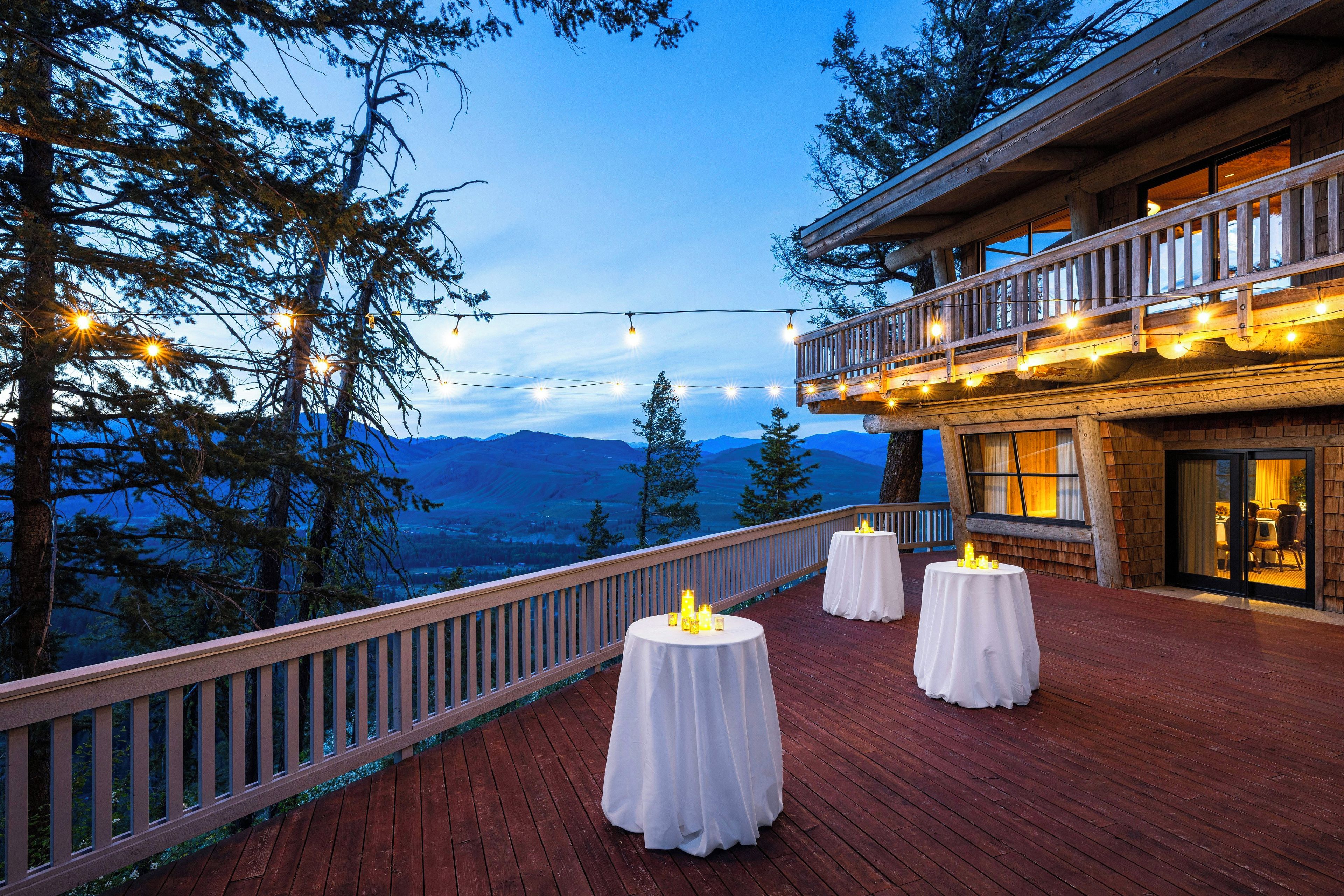 Um deck de madeira com mesas redondas, luzes de corda e vista para montanhas.