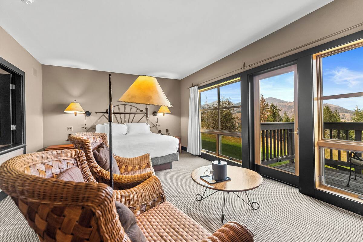 A hotel room with a large bed, wicker furniture, a round table, and a balcony with a view of the mountains.
