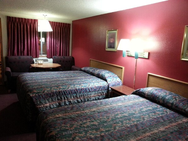 A hotel room with two beds, a small table, a chair, a lamp, and a window with curtains.