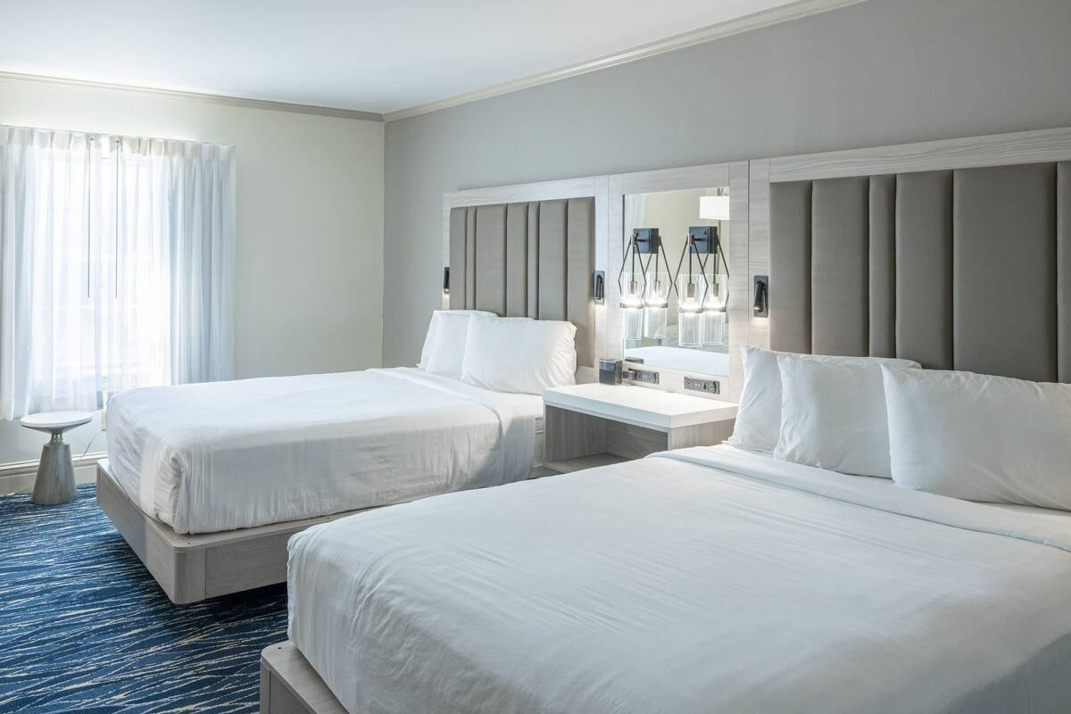 A hotel room with two beds, a bedside table, a nightstand, a mirror, and a window with curtains.