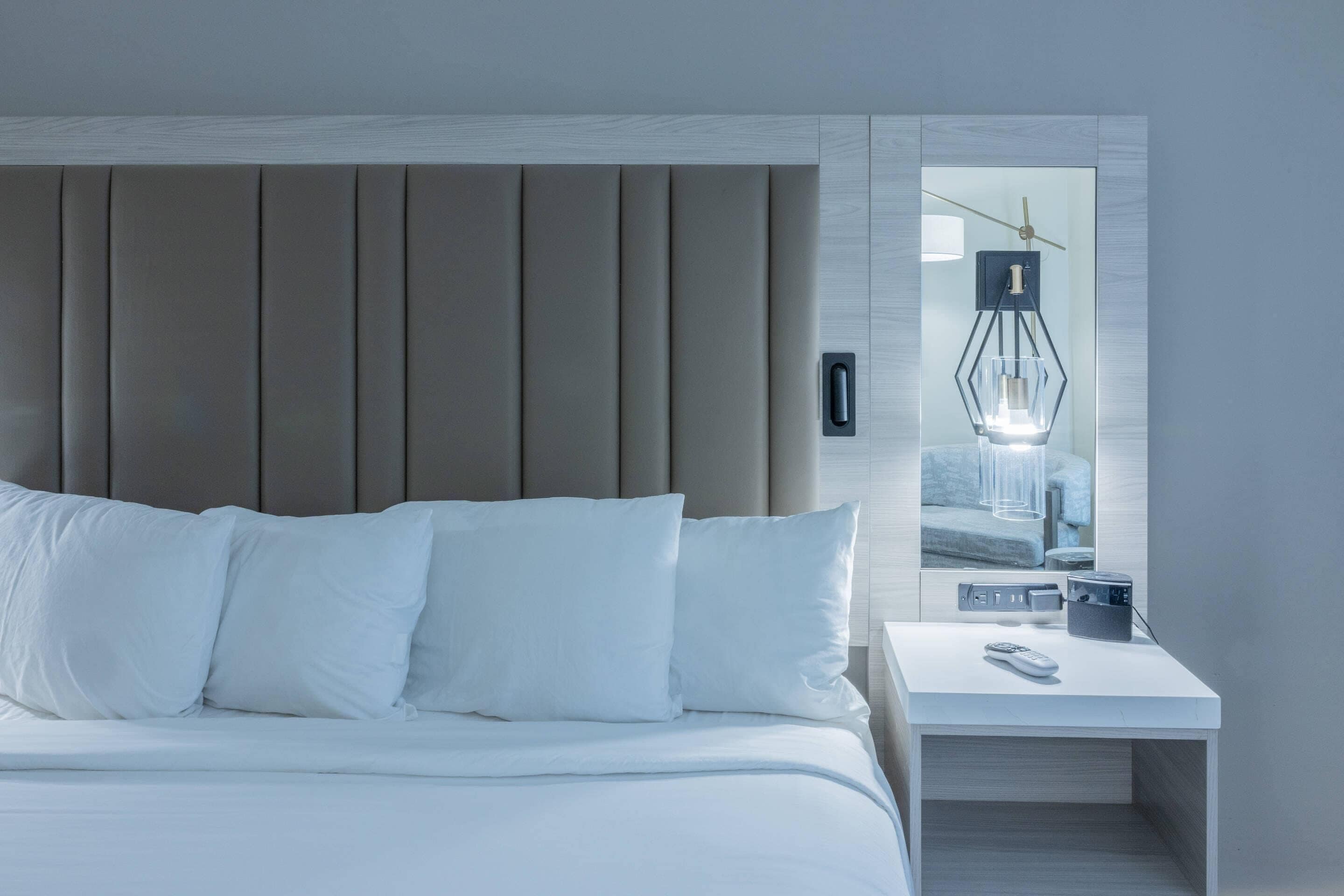 Una habitación de hotel moderna con una cama grande, una mesita de noche con lámpara y un espejo que refleja la habitación.