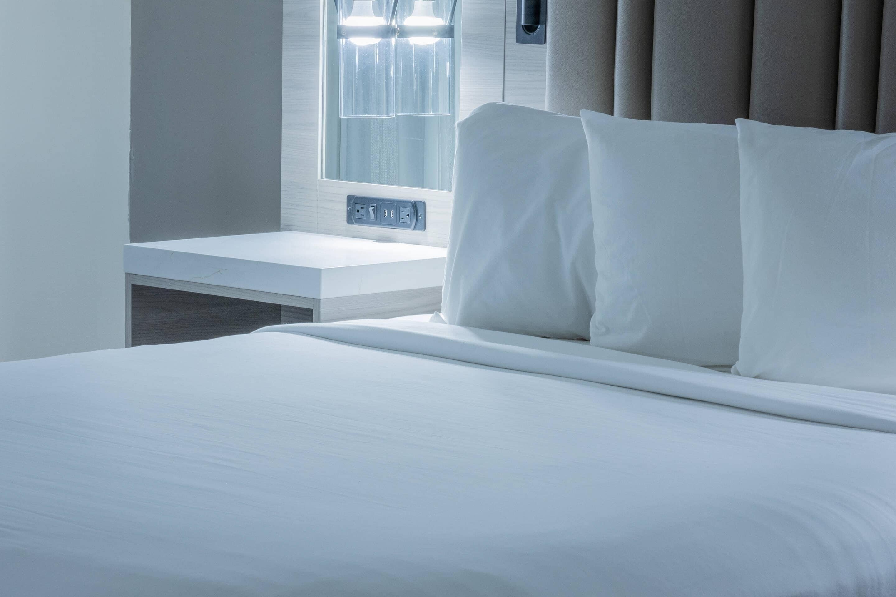 A neatly made bed with white linens, a bedside table with a power outlet, and a window with curtains.