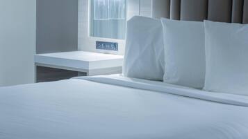 A neatly made bed with white linens, a bedside table with a power outlet, and a window with curtains.