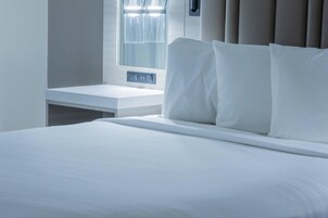 A neatly made bed with white linens, a bedside table with a power outlet, and a window with curtains.