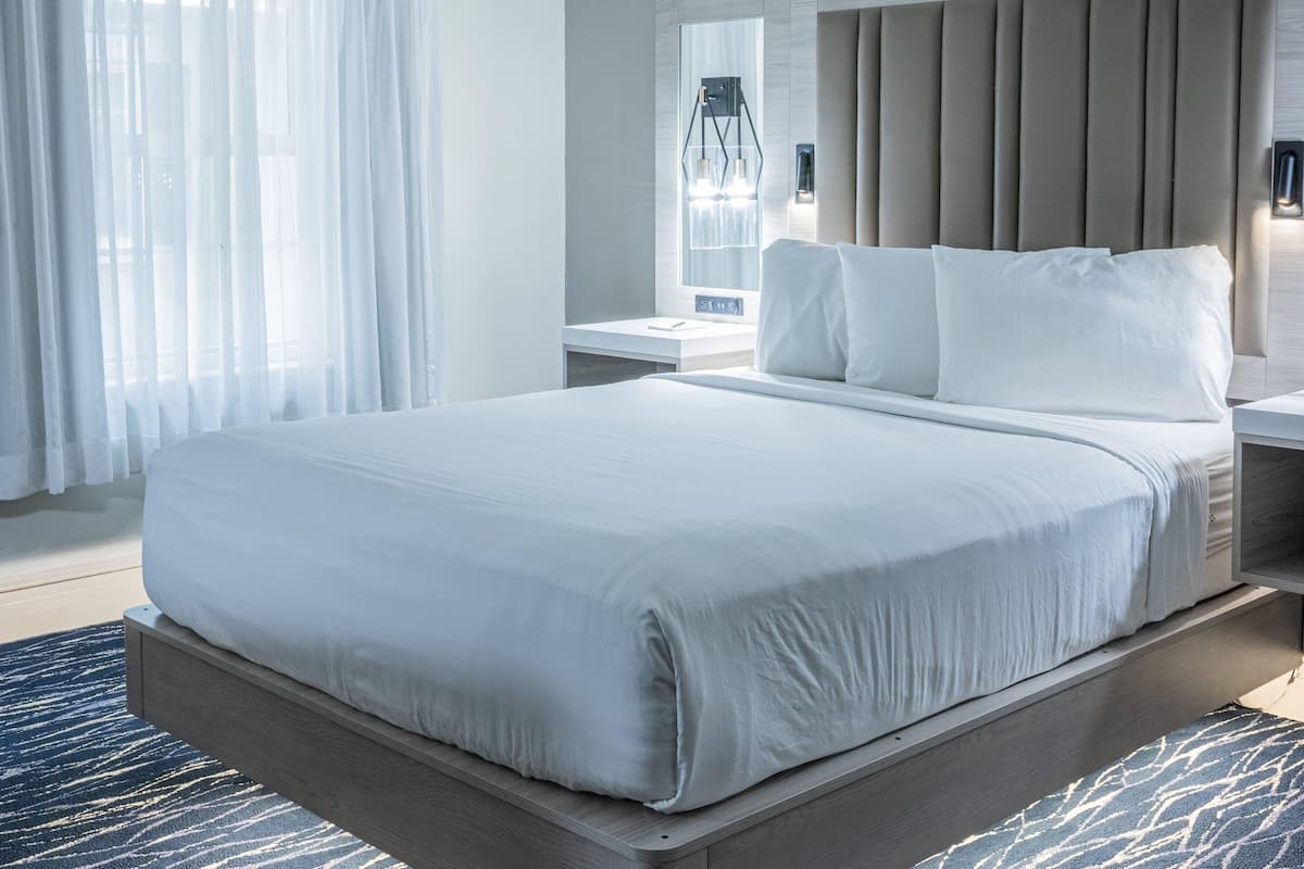 A modern hotel room with a large bed, a bedside table, a nightstand, and a window with sheer curtains.