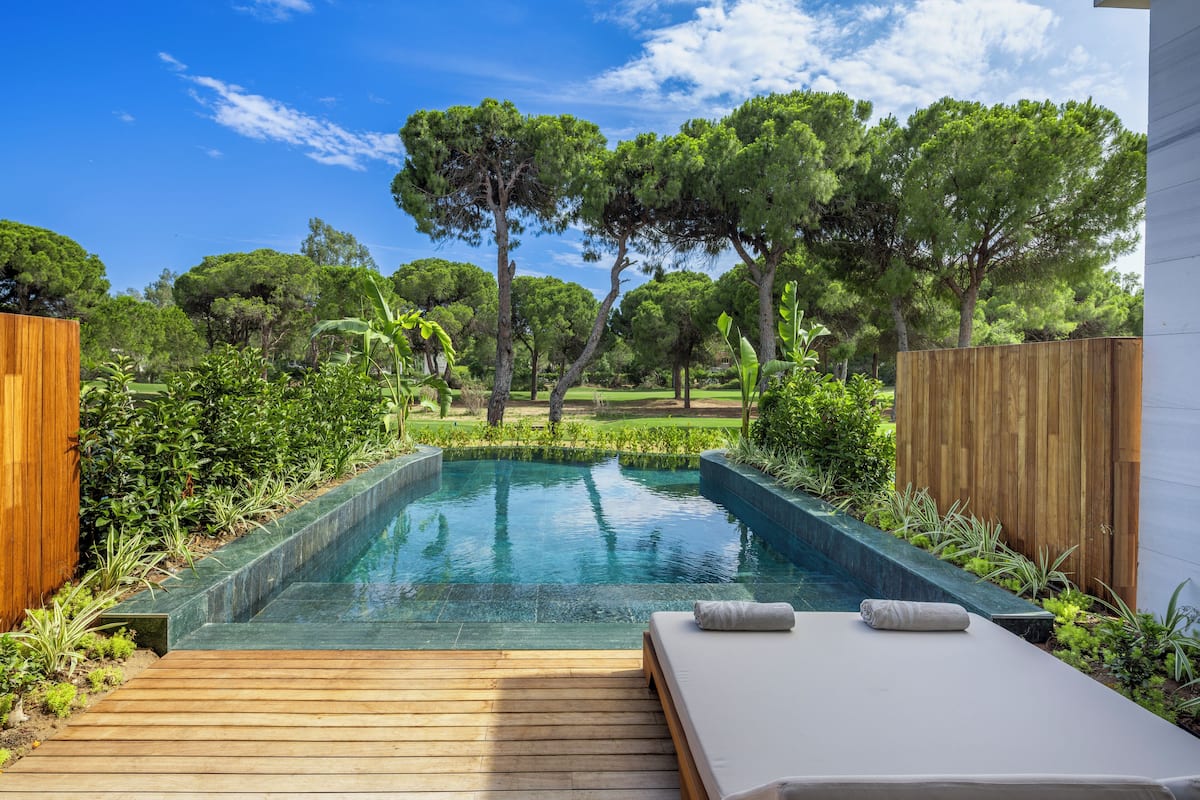 A rectangular swimming pool with a wooden deck in the foreground, surrounded by lush greenery and trees.