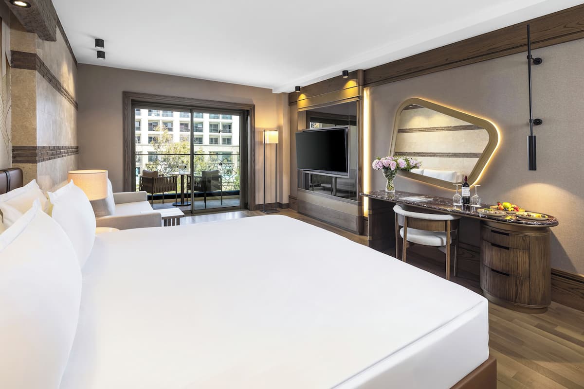 A modern hotel room with a large bed, a TV, a bathroom with a vanity, and a balcony with outdoor seating.