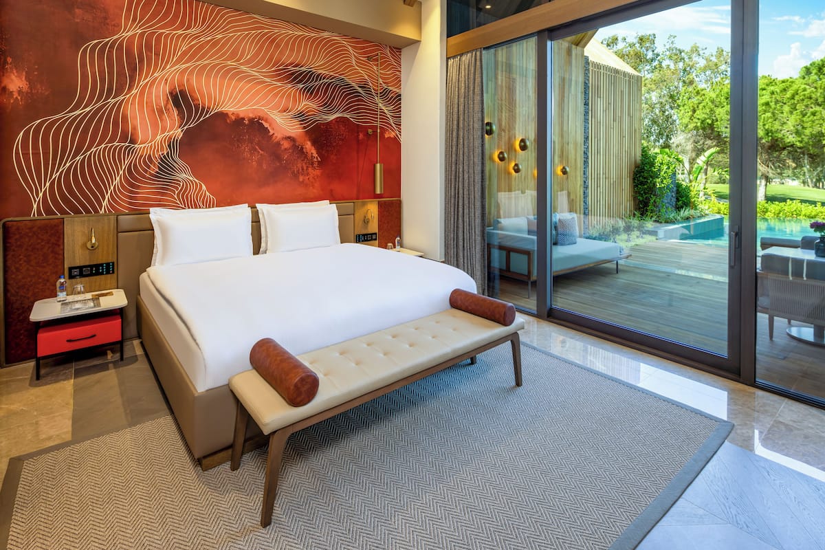 A modern hotel room with a large bed, a wall mural, a glass door leading to a balcony, and a wooden ceiling.