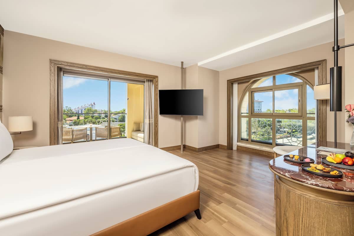 A hotel room with a large bed, a TV mounted on the wall, a dining area with a marble countertop, and large windows offering a view of the outdoors.