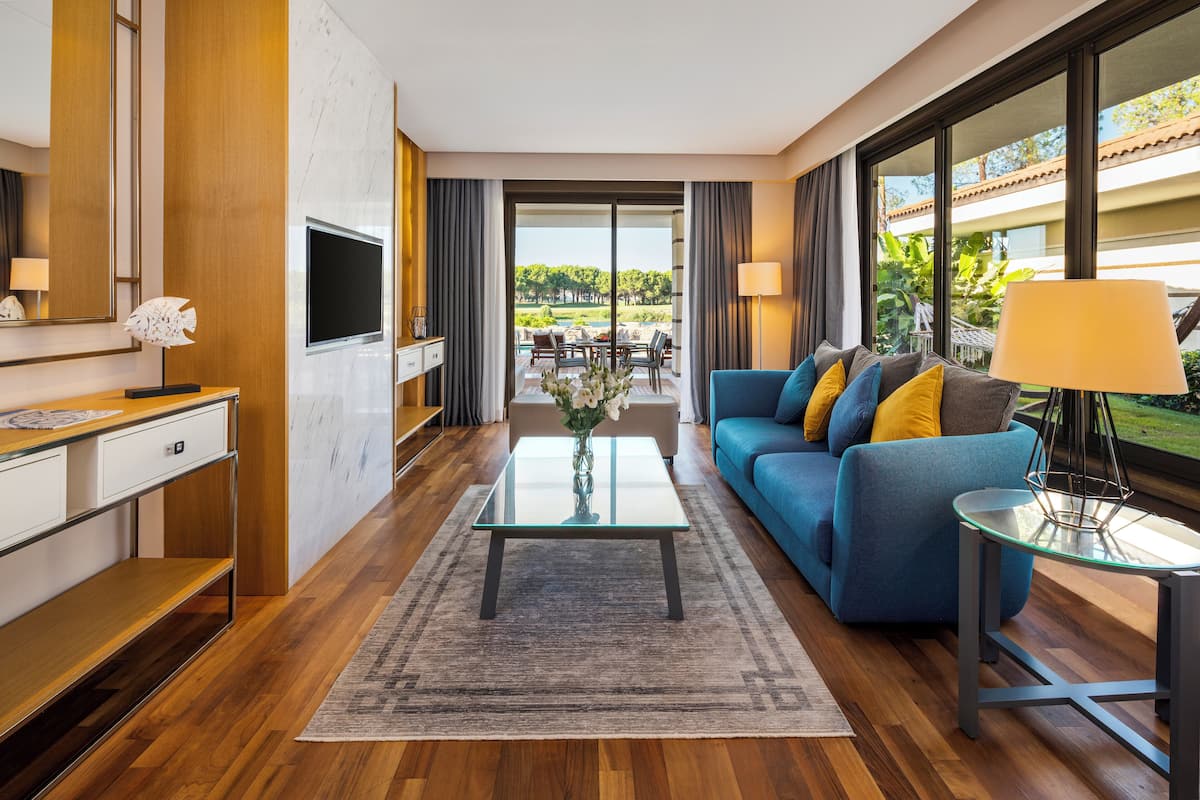 A modern hotel room with a wooden floor, a blue sofa, a glass coffee table, a TV, and a view of the outdoors.