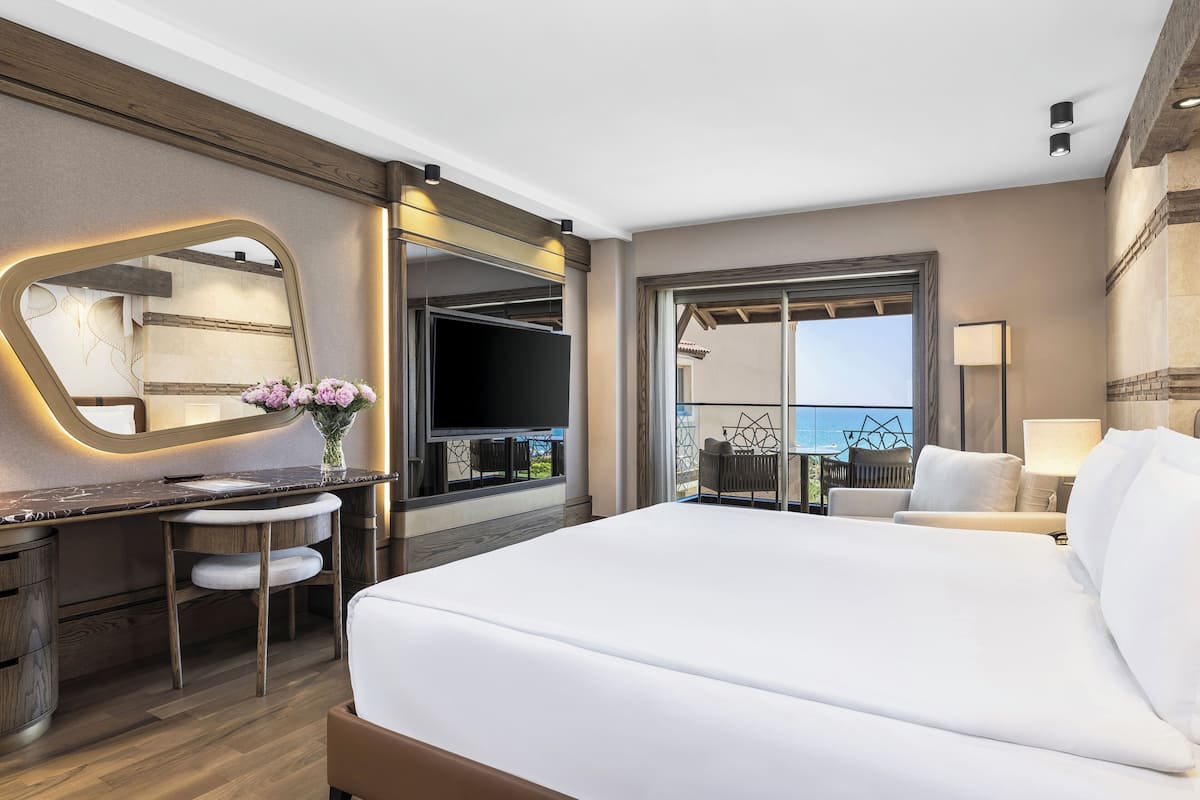A modern hotel room with a large bed, a desk with a chair, a TV, and a balcony with a view.