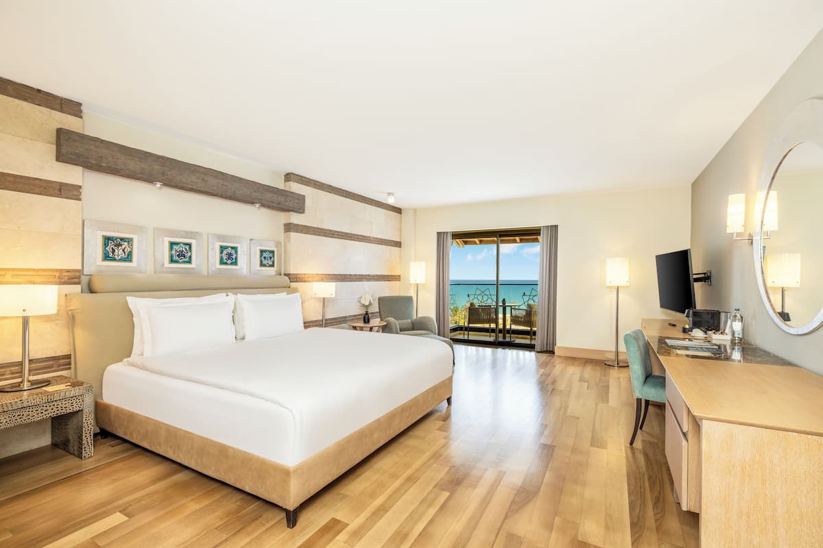 A hotel room with a large bed, a desk, a chair, a TV, and a balcony with a view of the sea.