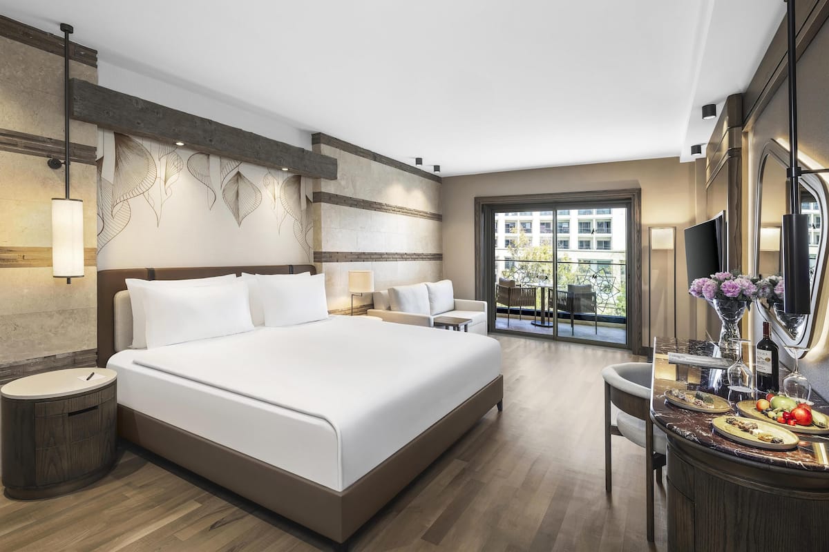 A modern hotel room with a large bed, a desk with a vase of flowers, and a view of the city through a large window.