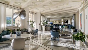 Un hall d’entrée spacieux avec un plafond haut, de grandes fenêtres et différents espaces de détente, notamment des canapés et des fauteuils.