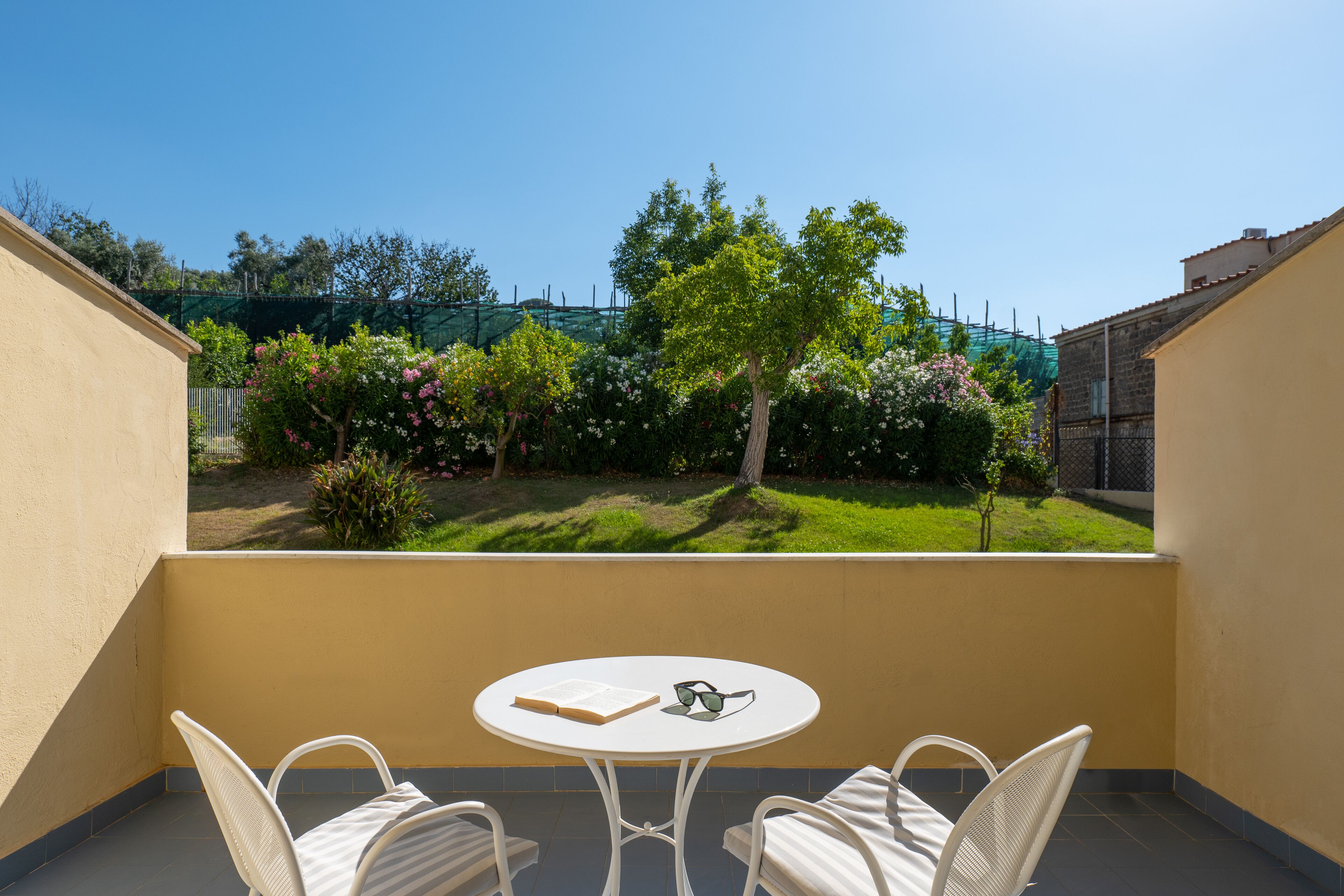 A balcony with a round table and two chairs, overlooking a garden with trees and flowers.
