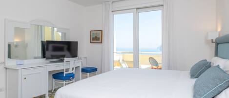 A hotel room with a bed, a desk with a computer, a chair, a television, and a window with a view of the sea.