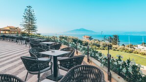 Un espace extérieur aménagé avec des chaises et des tables en osier, offrant une vue sur un paysage côtier avec une montagne à l’horizon.