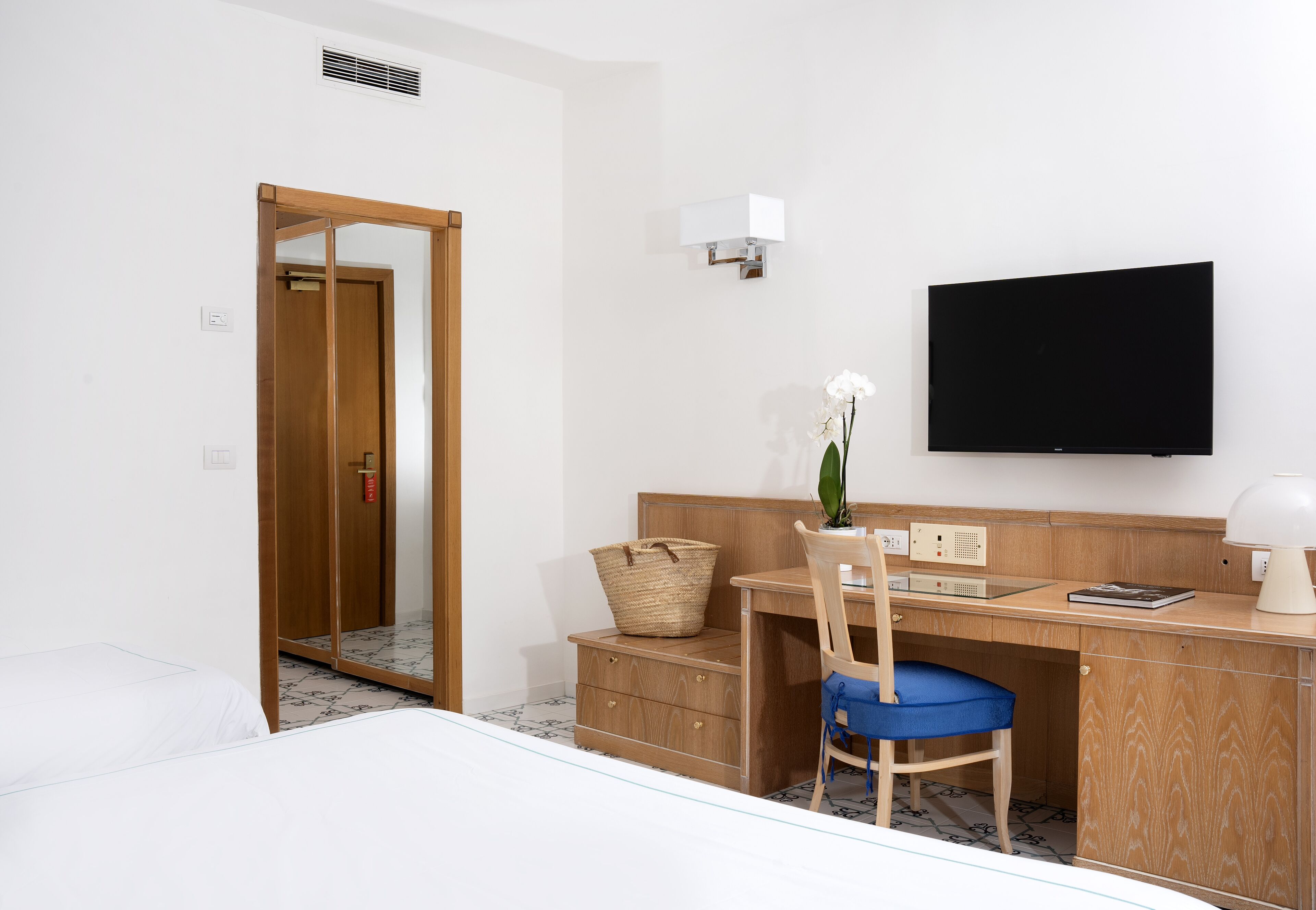 Hotellihuone, jossa on sänky, työpöytä tuolilla, televisio ja pieni ruukkukasvi.