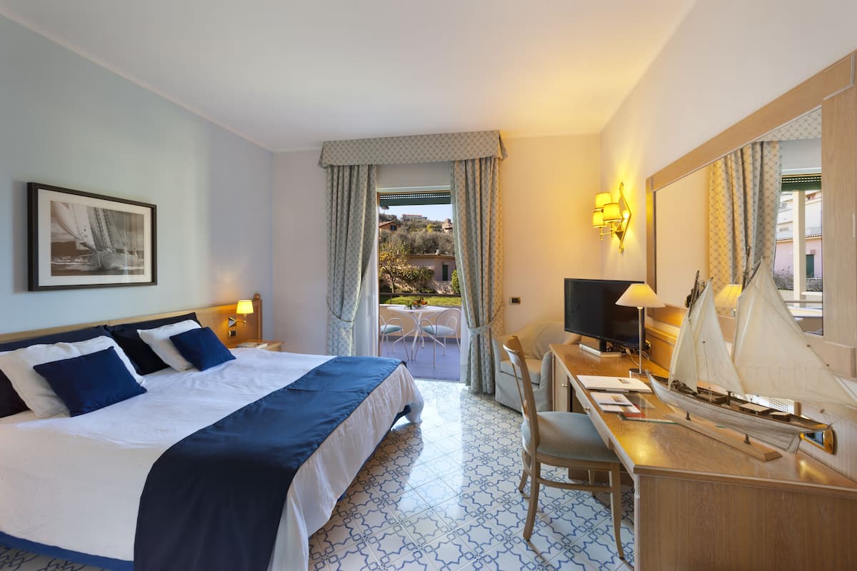Habitación de hotel con cama, escritorio, silla, un modelo de velero, televisor y balcón con mobiliario exterior.