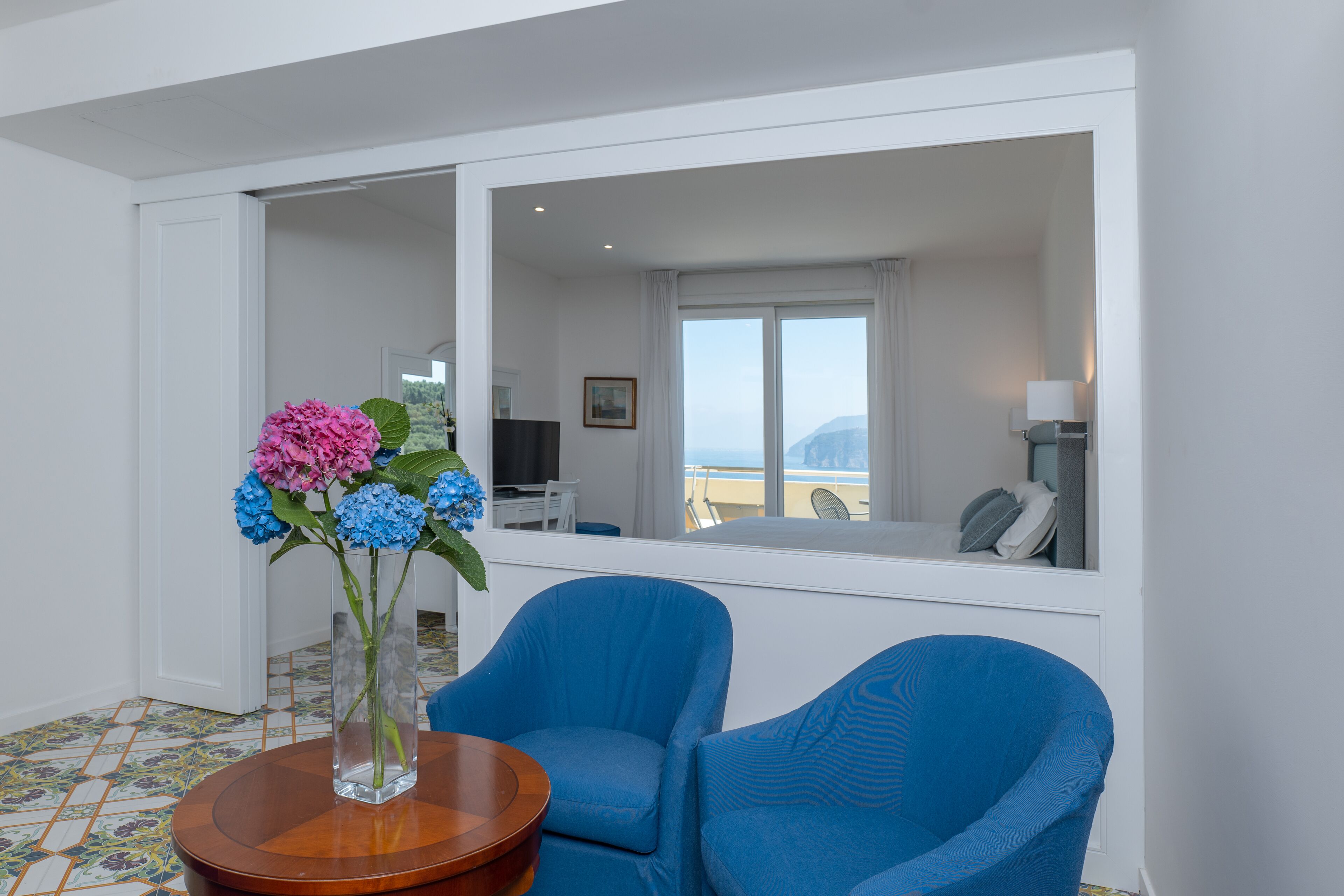 A room with a large window, a mirror, a vase with flowers, and two blue chairs.