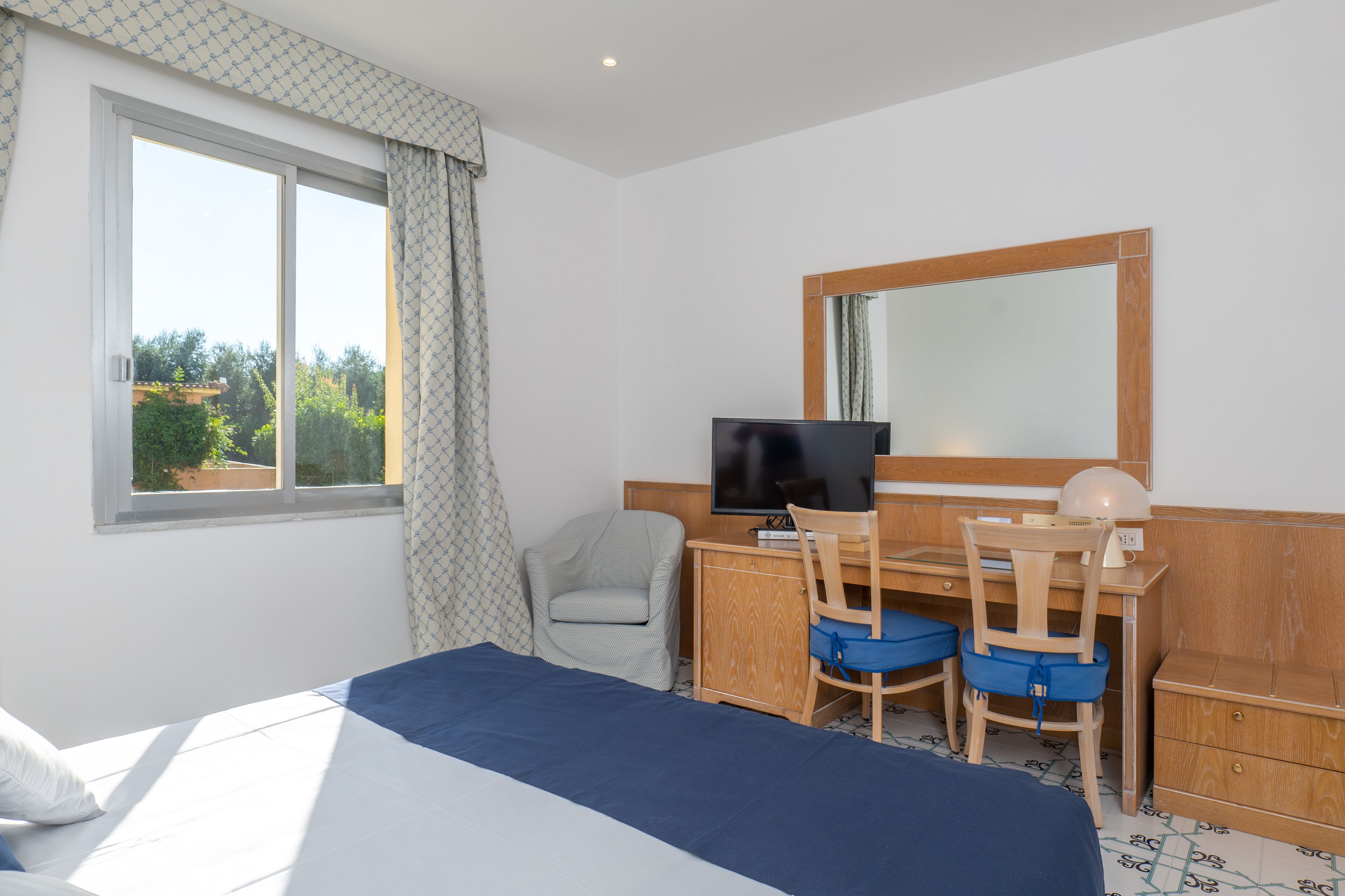 A hotel room with a bed, a desk with a computer and chair, a television, and a window with curtains.