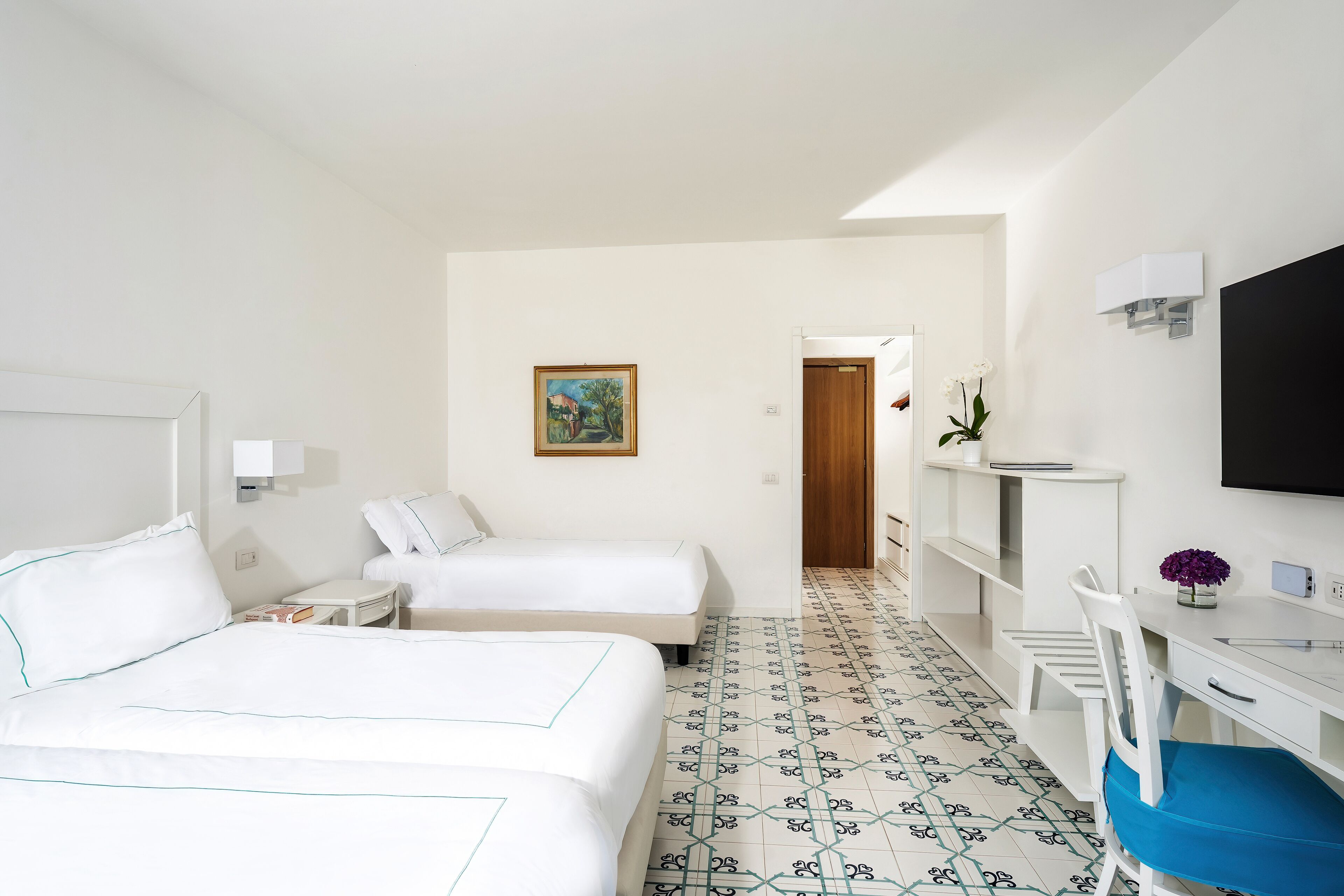 A hotel room with two beds, a desk, a chair, a TV, and a painting on the wall.