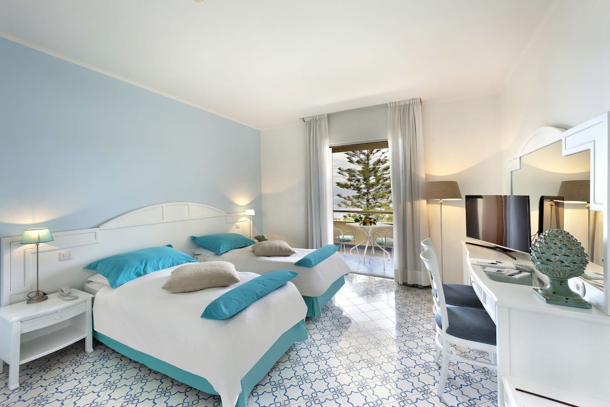 Habitación de hotel con dos camas, un escritorio, una silla, un televisor y un balcón con vista a un árbol.