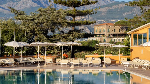 Un espace piscine avec des chaises longues et des parasols, un paysage montagneux en toile de fond et un bâtiment doté d’un balcon.