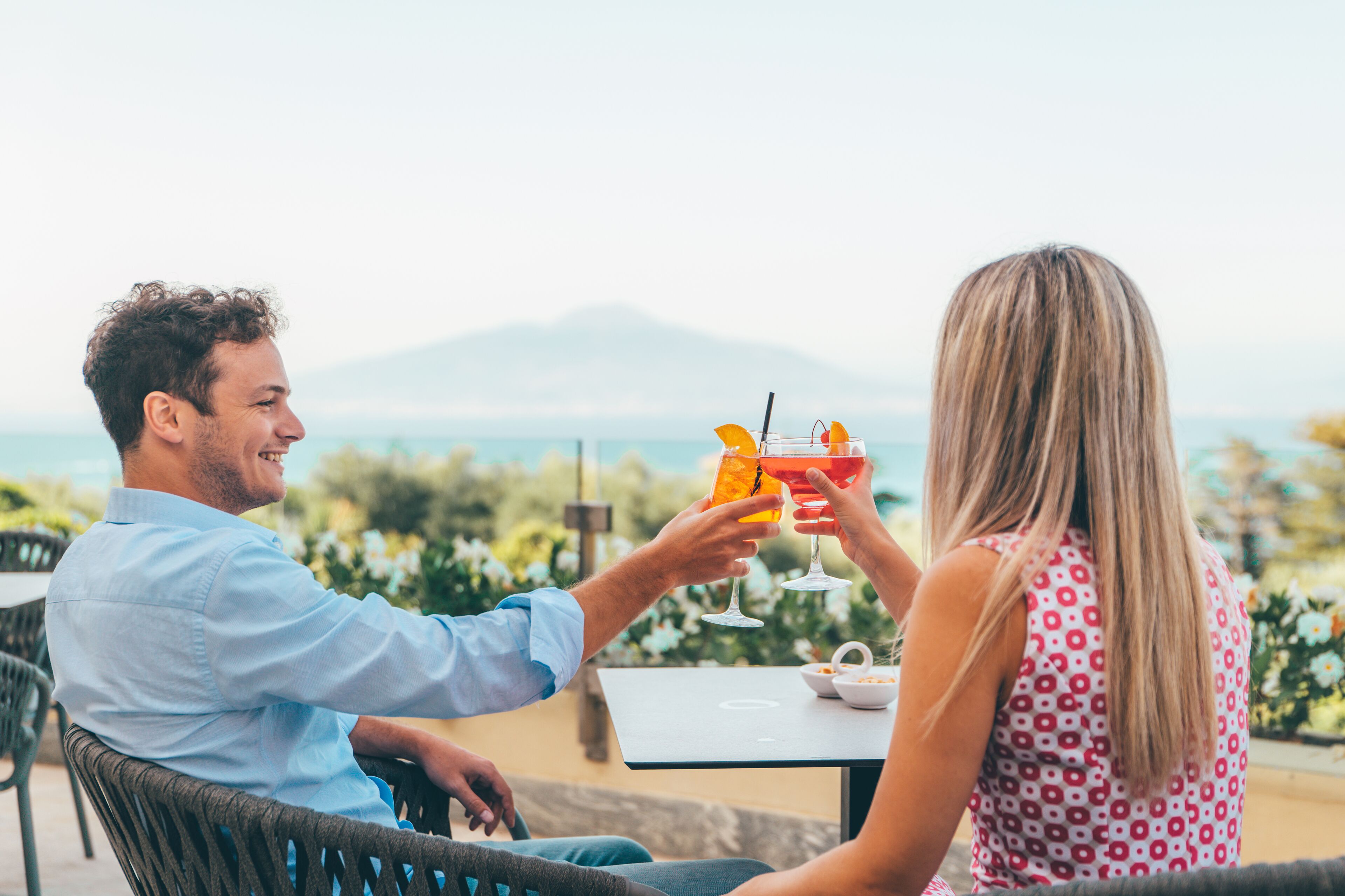 A couple toasting with drinks at an outdoor table with a view of the sea and mountains.