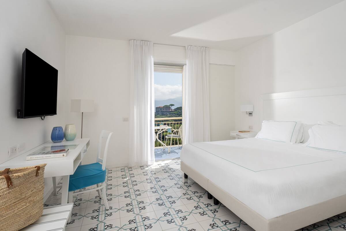 Habitación de hotel con cama, escritorio, silla, televisor y balcón con vistas.