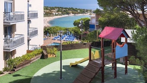 Un parque infantil con tobogán y estructura de escalada, rodeado de una zona de piscina con sillones de descanso y vistas a una playa.