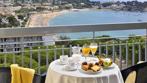 Una mesa en el balcón con alimentos para el desayuno, una vista panorámica de una playa y el océano, y un edificio de hotel al fondo.