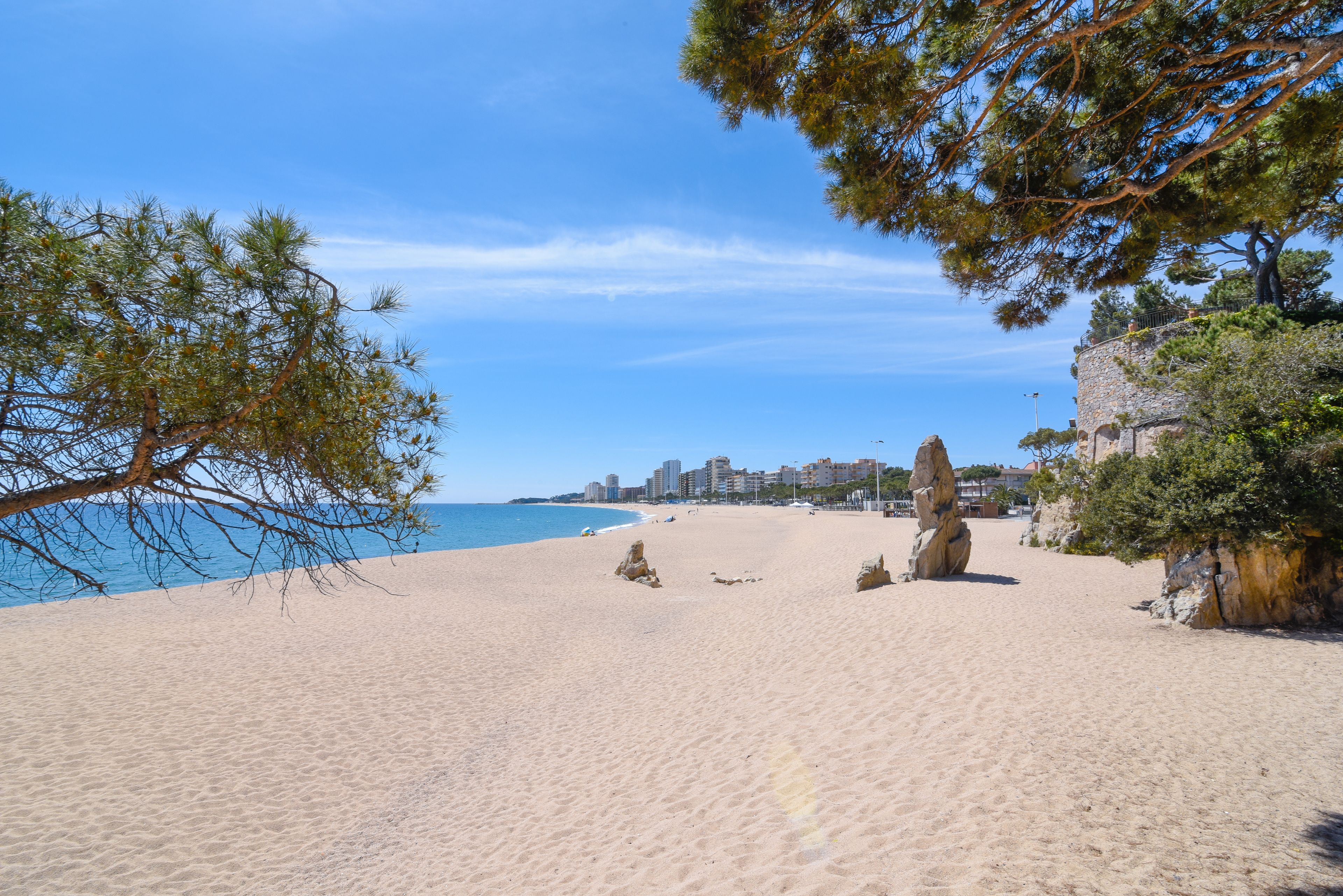 A sandy beach with a clear blue sky, a rocky outcrop, and a distant city skyline.