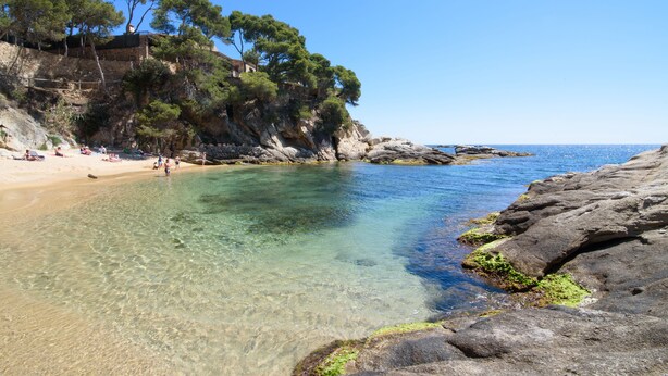 A coastal beach with clear water, rocky shore, and a stone building on a cliff.