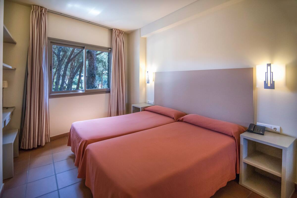 Ein Zimmer mit zwei Betten, einem Fenster mit Vorhängen, einem Nachttisch mit Telefon und einer Wandleuchte.