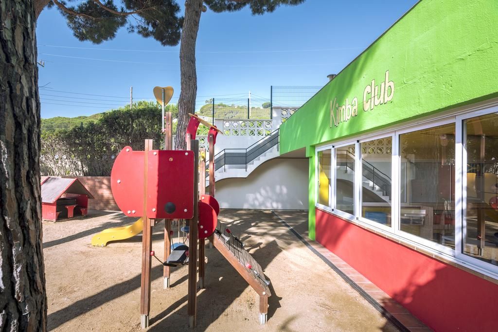 A children's play area with a red climbing structure and a slide, adjacent to a building with a green facade and the sign 'Kimba Club'.
