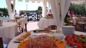 Un área de comedor al aire libre con mesas preparadas para una comida, mostrando una variedad de alimentos, incluyendo frutas, verduras y carnes.