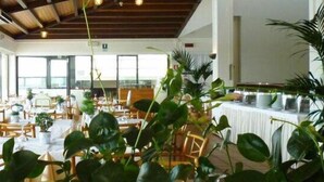Un espacio interior amplio con varias mesas y sillas, ventanas grandes y plantas en macetas.