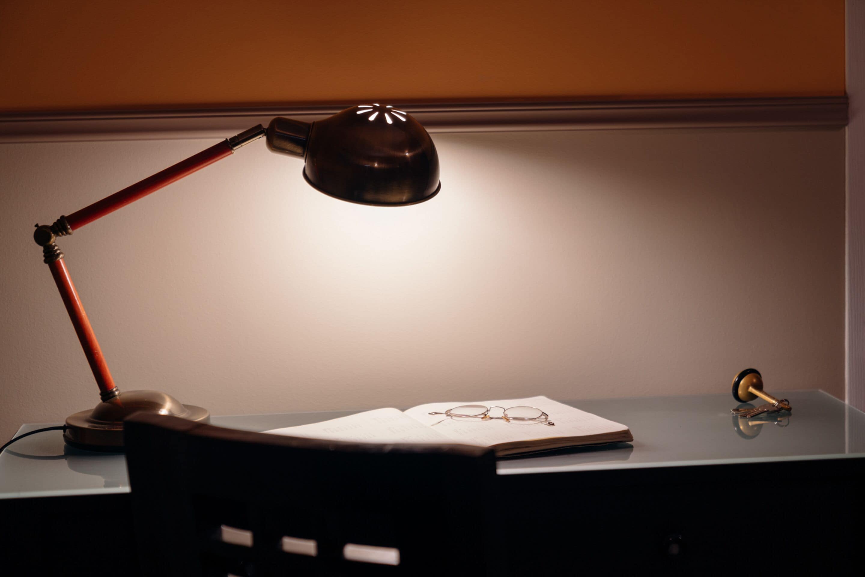 Una scrivania con una lampada, un libro aperto e un paio di occhiali.