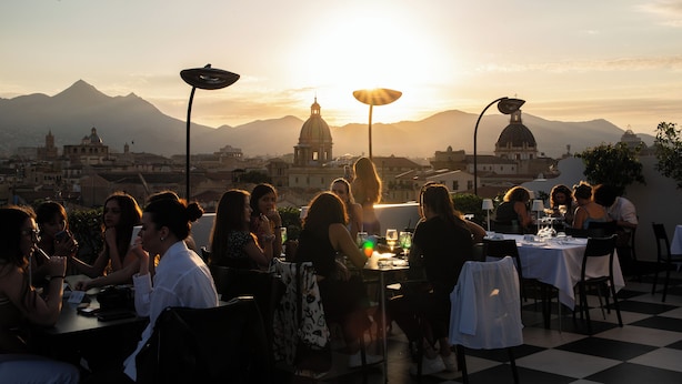 Un área de comedor en la azotea con mesas y sillas, con vistas a un paisaje urbano con edificios de cúpulas y montañas al fondo.