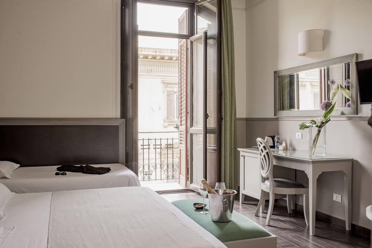 A hotel room with two beds, a desk with a chair, a minibar, and a large window with a view of a building.