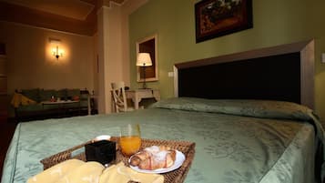 Habitación de hotel con cama, mesita de noche, lámpara, cuadro enmarcado en la pared y bandeja de desayuno con cruasanes y jugo de naranja.